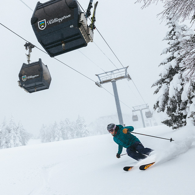 Skier carving through fresh snow beneath a gondola on a snowy mountain.
